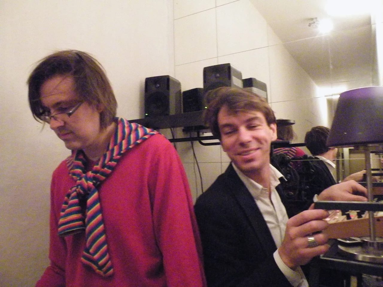 Two men at a house party — one in a pink jumper and striped scarf looking down, the other in a dark jacket grinning at the camera — with studio monitors on a shelf behind them.