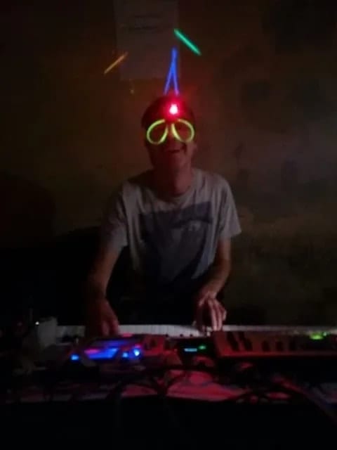 Playing a synthesizer in a dark room, wearing glowstick glasses and a light-up headband with a red LED, the controllers below lit blue and pink.
