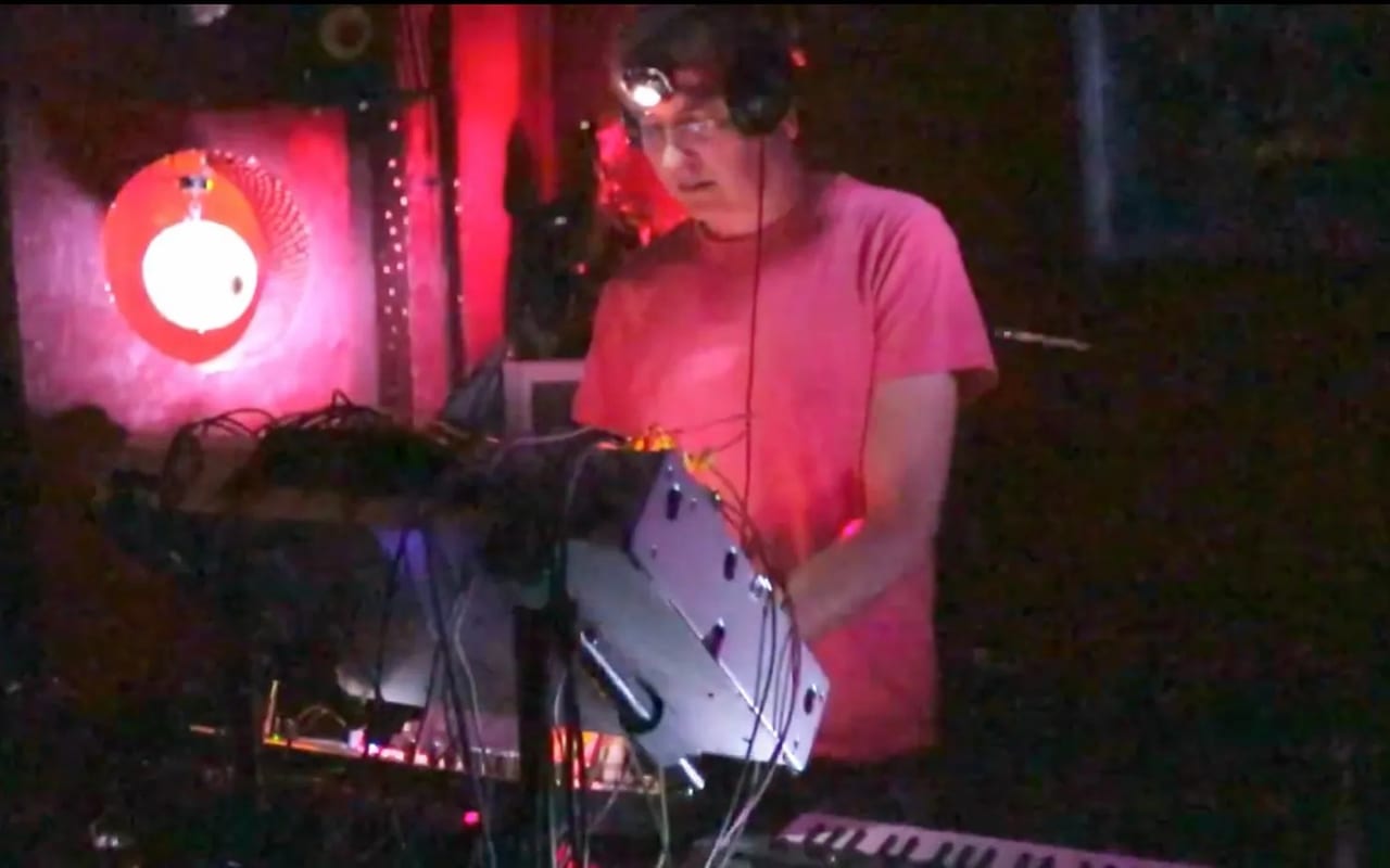 Playing a patched modular rig in a pink t-shirt and headphones, under red stage lighting.