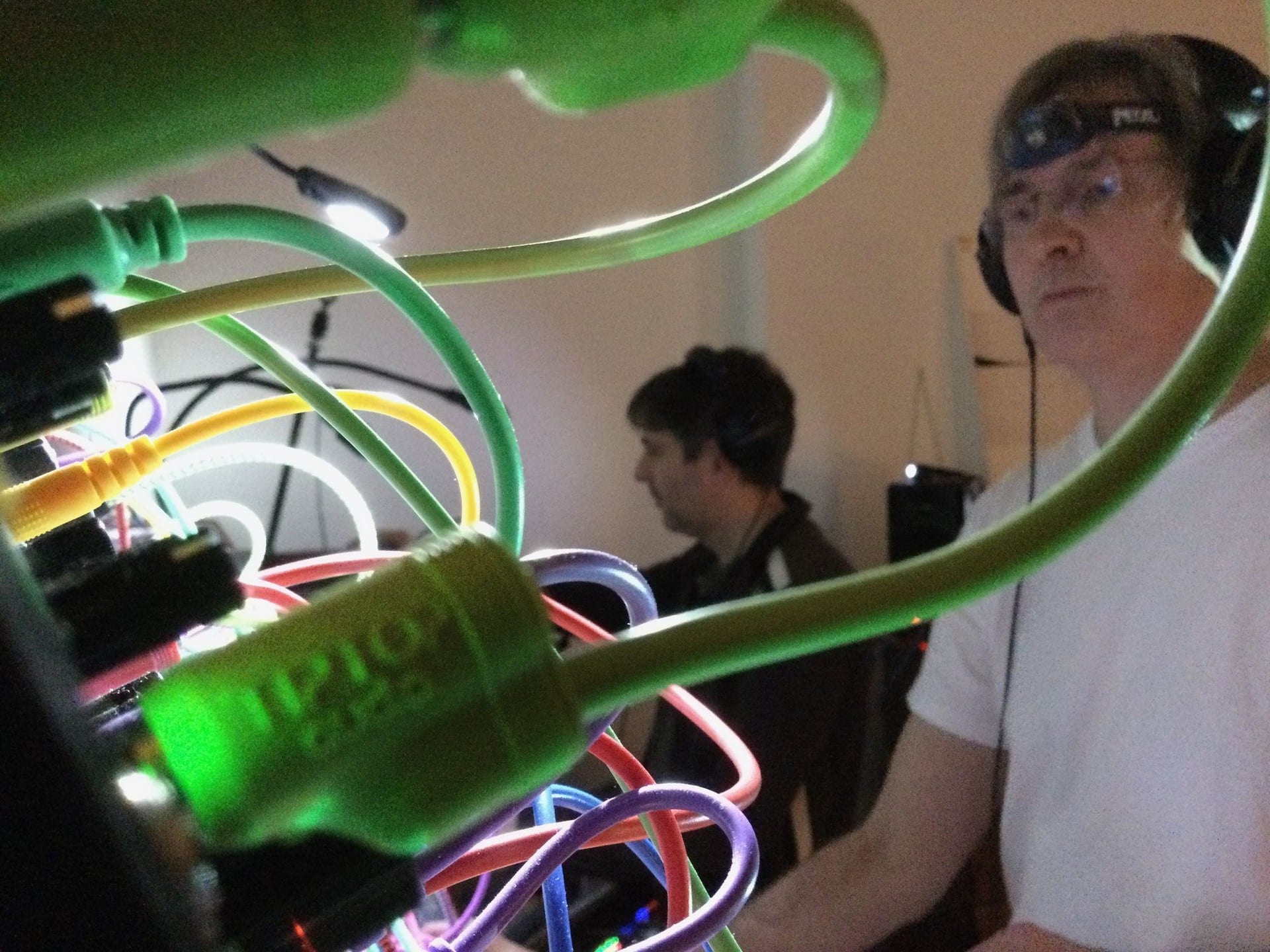 Close-up through a tangle of green and multicoloured patch cables on a Eurorack synth, with two people playing behind.