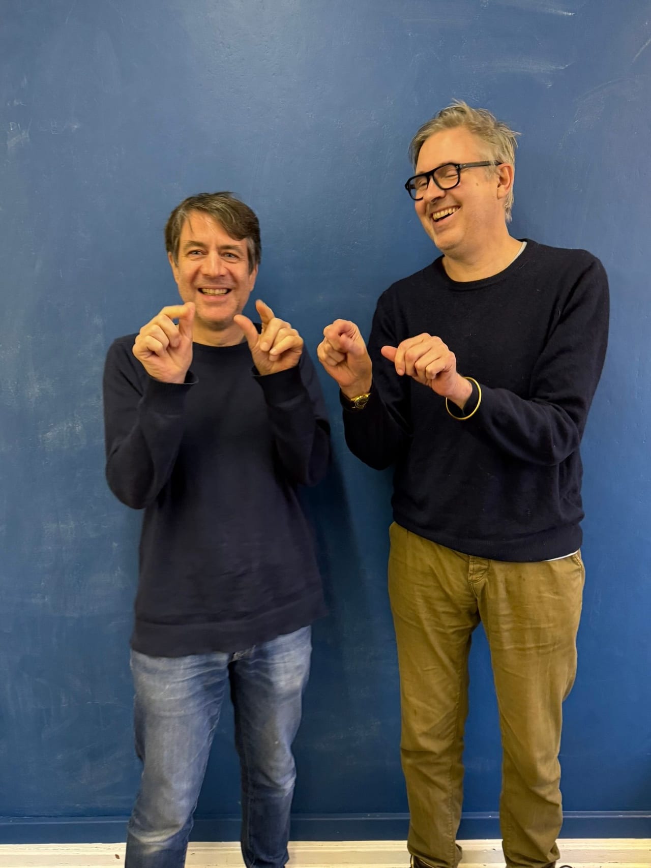 Two members of 3rd Party Influence against a blue wall in dark jumpers, laughing and pointing at each other.