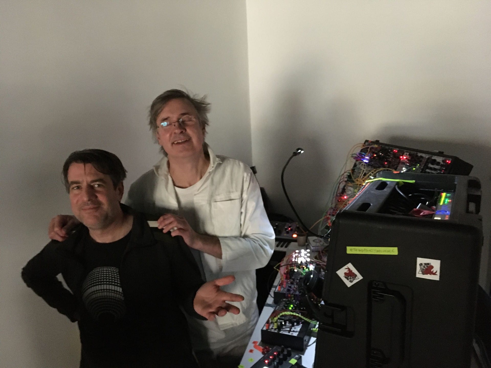 Two performers beside a Eurorack case covered in lit modules, one with an arm around the other.