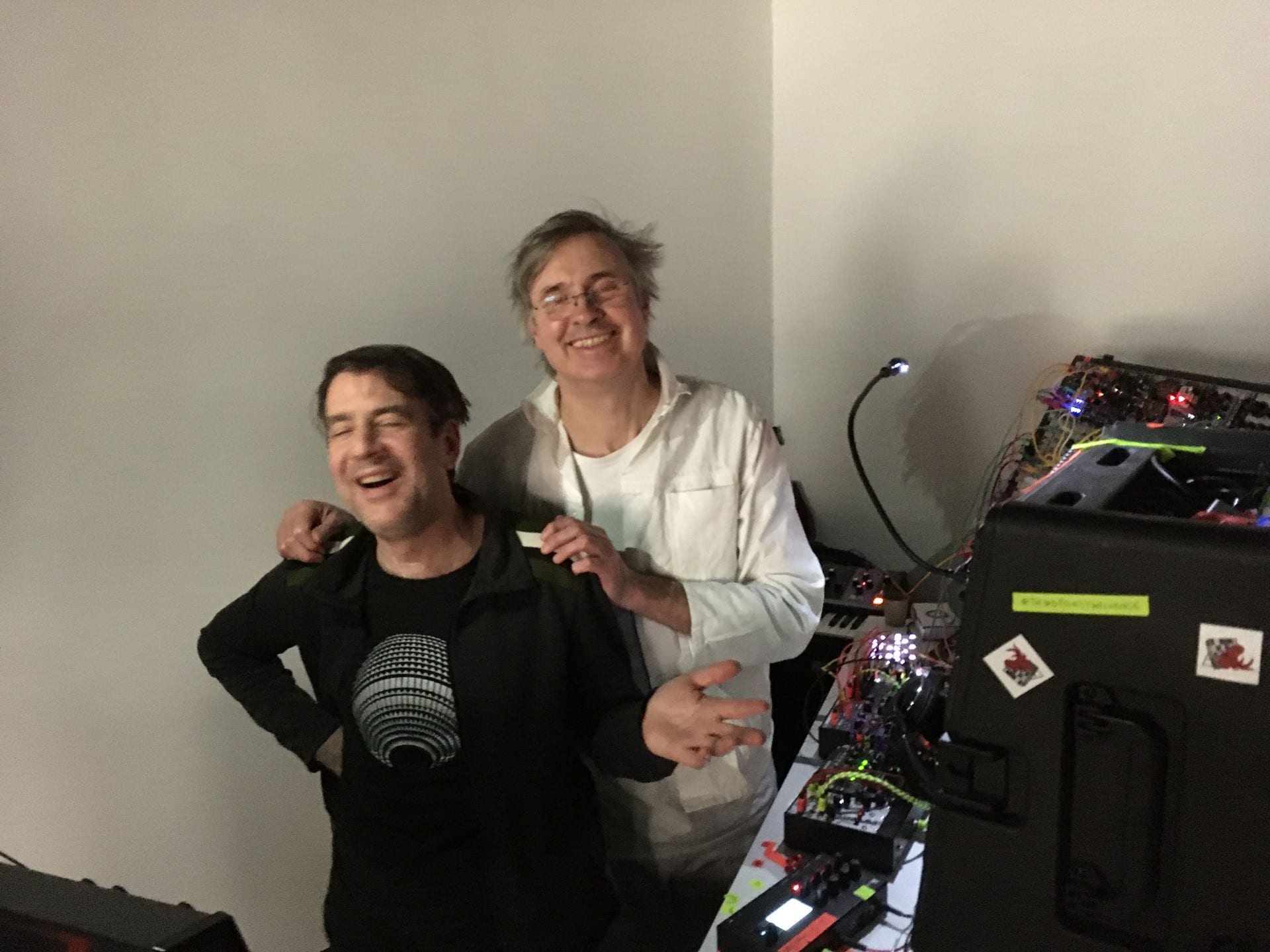 Two performers laughing together beside a Eurorack case covered in lit modules.