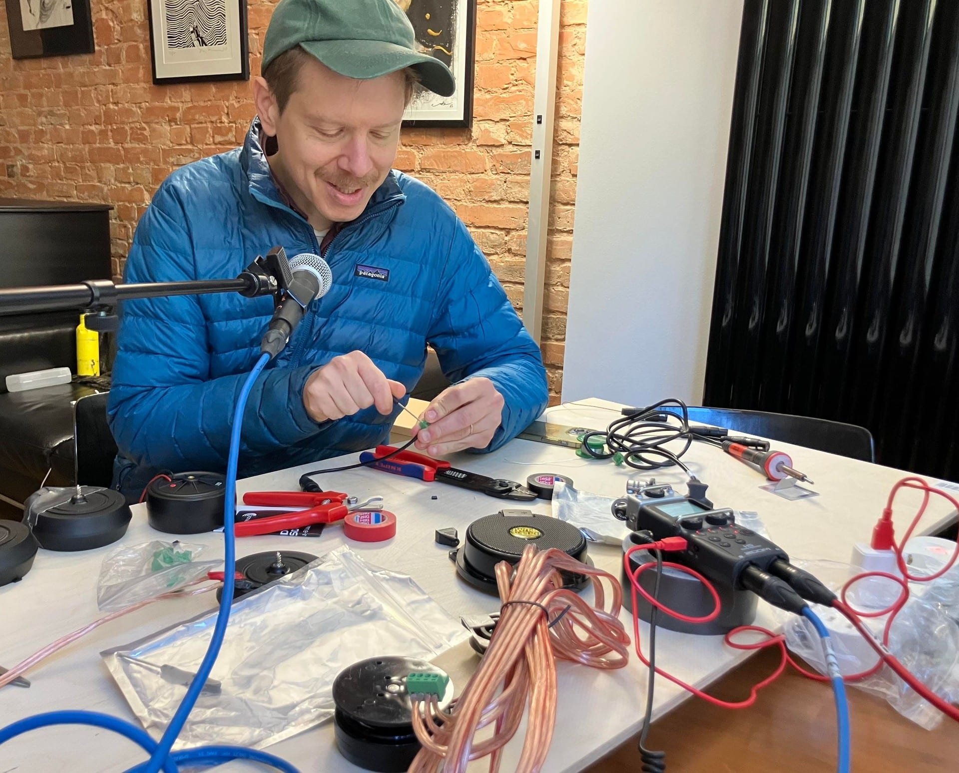 A man in a blue puffer jacket and cap assembling electronics at a table, among coils of copper wire, pliers and a multimeter.