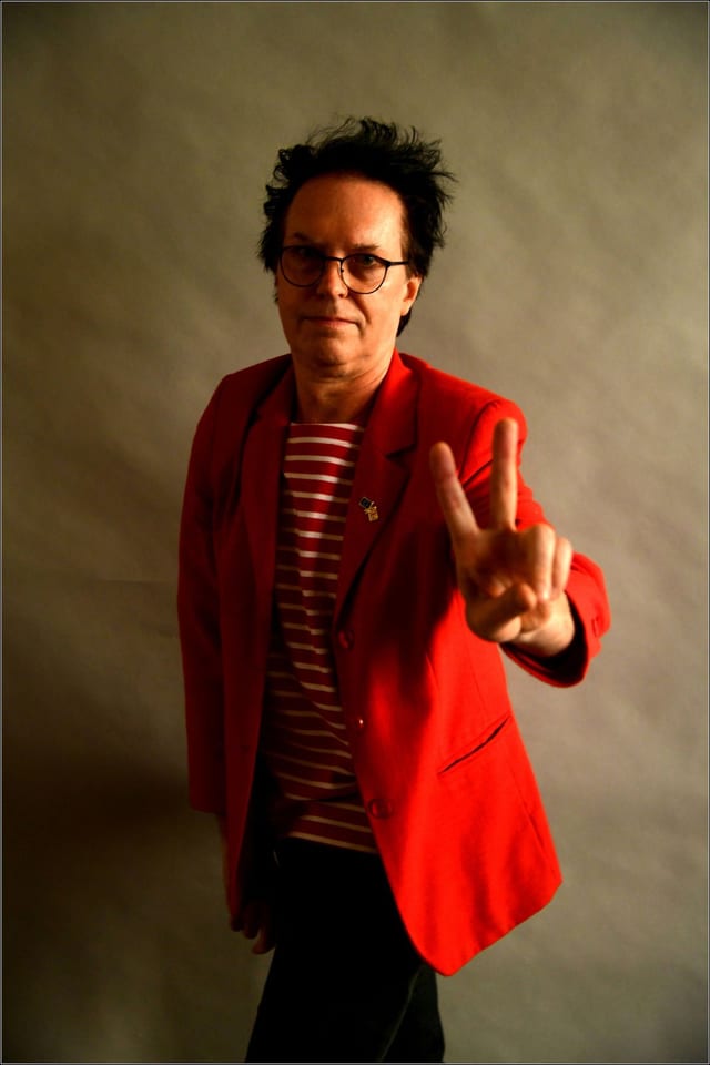 A man with spiked hair and round glasses in a bright red blazer over a striped top, making a V sign at the camera.