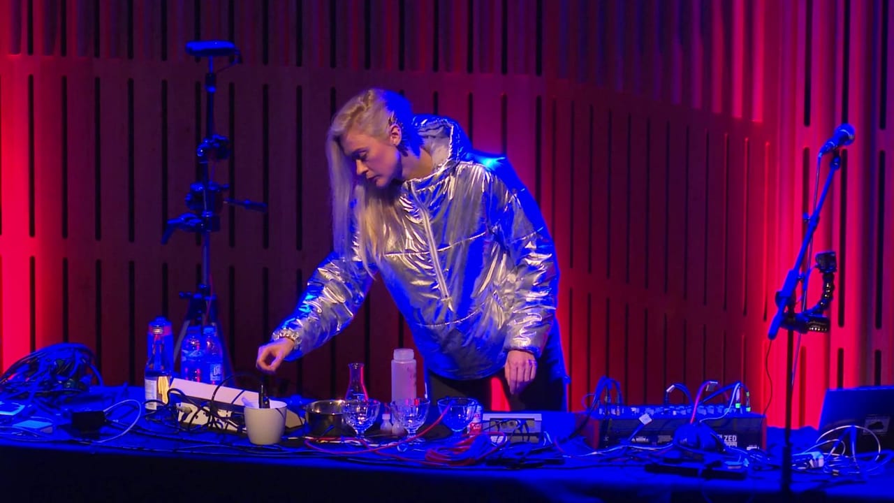 A performer with platinum hair in a silver metallic jacket, leaning over a table of laboratory glassware and electronics on a red-lit stage.