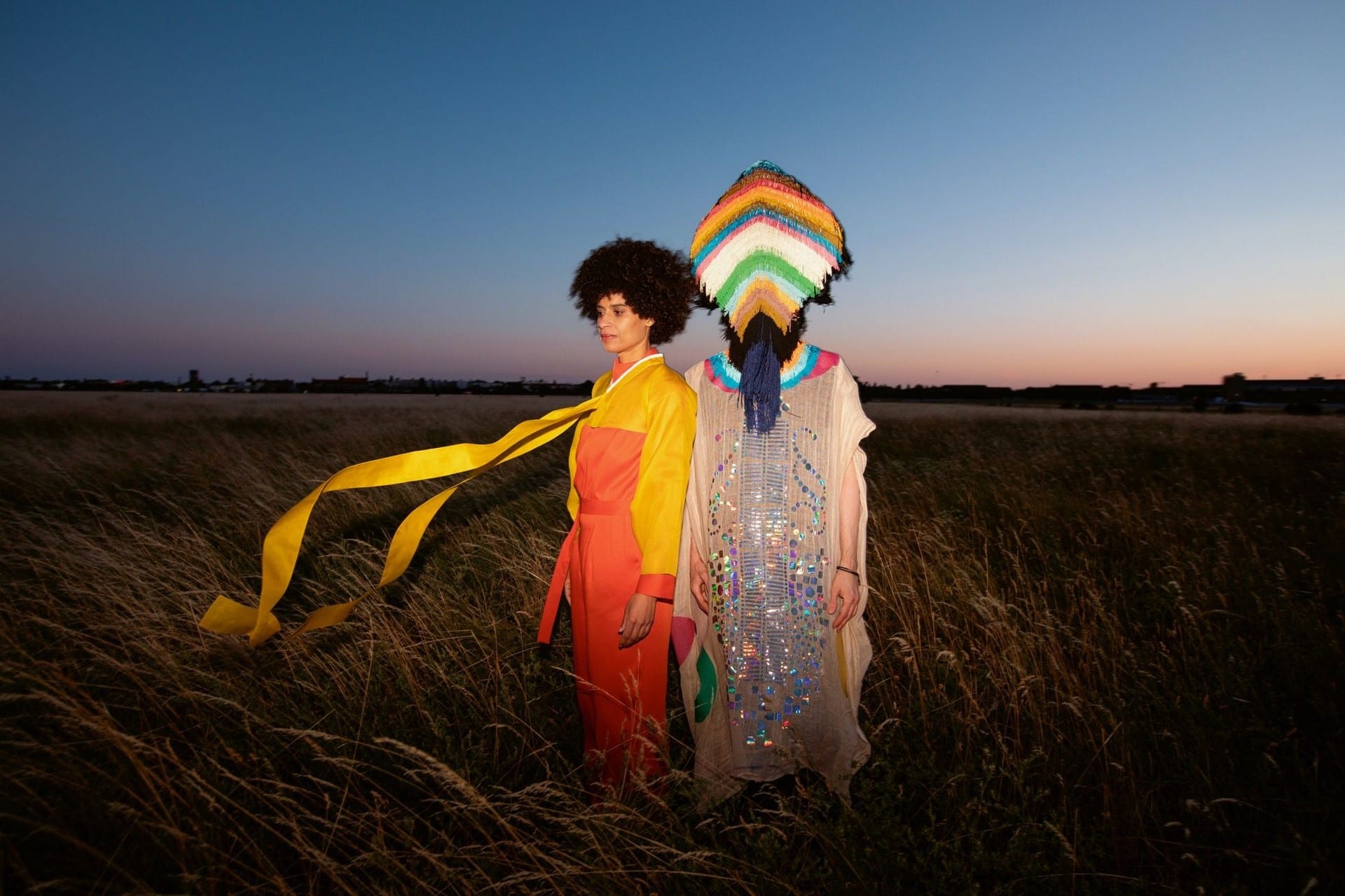 Two figures standing in tall grass at dusk: one in red and yellow with a long ribbon streaming in the wind, the other in a pale sequinned robe and a rainbow fringed headdress covering the face.