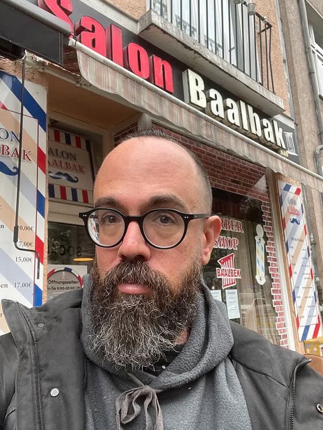 A bearded man in round glasses and a grey hooded jacket, photographed outside the Salon Baalbak barber shop in Berlin.