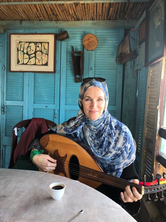 A woman in a blue patterned headscarf playing an oud at a table, in a room with turquoise wooden shutters, a small cup of coffee beside her.
