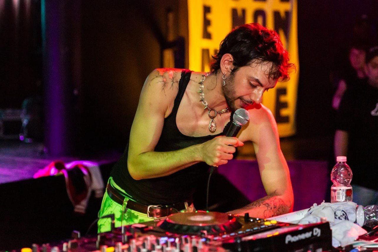 A performer in a black vest and chain necklace singing into a microphone over DJ decks, lit pink and yellow.