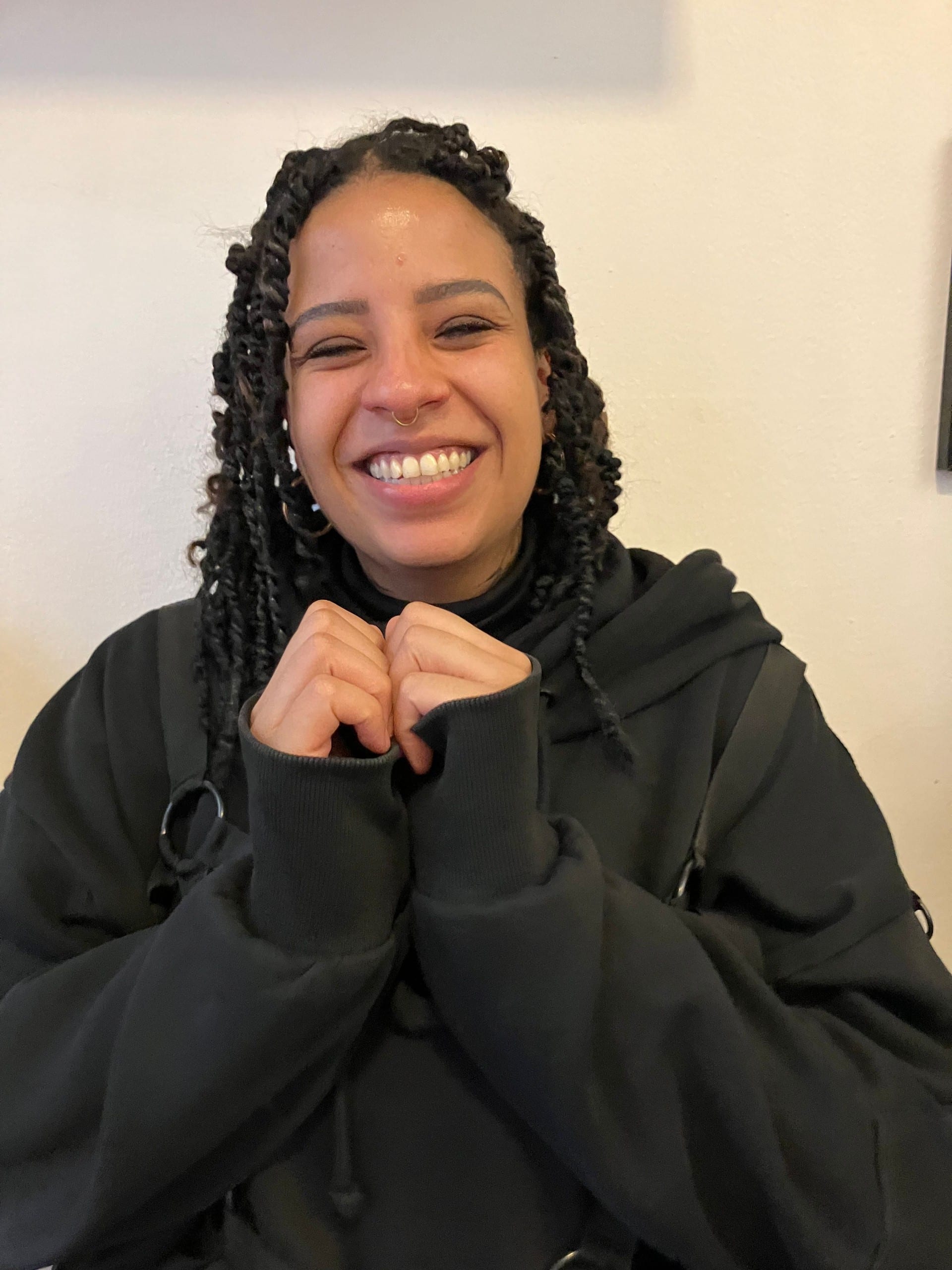A smiling person with braided hair in a black hoodie, hands drawn up to their chin.